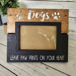 May include: A rectangular photo frame featuring a black wooden base with the white text LEAVE PAW PRINTS ON YOUR HEART and a copper-toned metal top panel with cutouts of the word Dogs and two paw prints, secured by two visible screws.