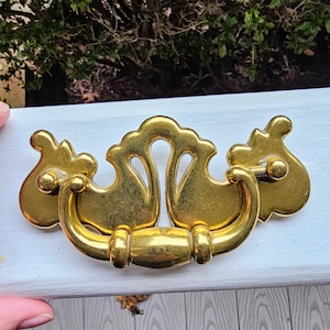 May include: A gold-toned drawer pull with a decorative design. The pull features a central handle with a curved design and two side pieces. The hardware is mounted on a white surface.