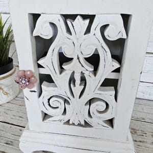 May include: White wooden cabinet with a carved design on the front. The cabinet has a distressed finish and a pink flower embellishment.