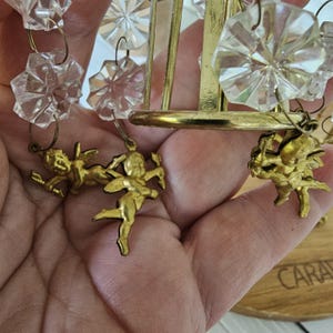 May include: Close-up of a vintage chandelier with clear crystal accents and gold-colored metal framework. The chandelier features small, gold-colored cherub figurines hanging from the frame. The crystals are faceted and reflect light.
