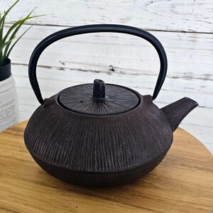 May include: A dark brown cast iron teapot with a black handle and lid. The teapot has a textured, ribbed surface and a spout. The handle arches over the top. The teapot sits on a wooden surface.