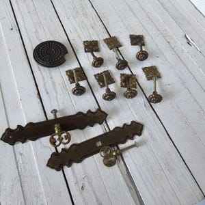 May include: An assortment of antique brass hardware, including decorative pulls, knobs, and a circular plate. The pieces have an aged patina and ornate floral and keyhole designs, set against a white wooden surface.