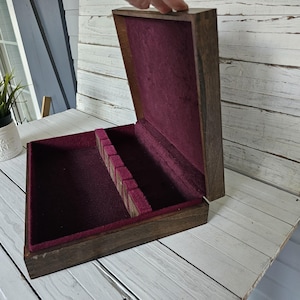 May include: Open wooden box with a dark stain, lined with burgundy fabric. The box has compartments for storage and is partially open, revealing the interior. The box is sitting on a white wooden surface.