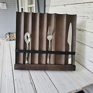 May include: A wooden cutlery holder with a wavy design, holding a spoon, fork, and knife. The holder is dark brown with a black band securing the silverware. The silverware is made of shiny metal.