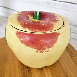 May include: A ceramic apple-shaped container with a lid. The container is primarily yellow with red accents, mimicking the appearance of a ripe apple. The lid features a green stem. The container is sitting on a wooden surface.