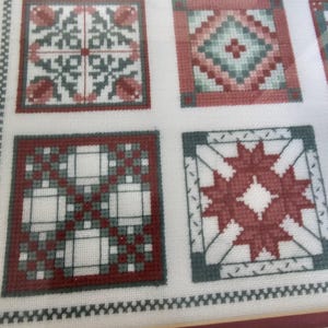 May include: Close-up of a cross-stitch sampler featuring four square designs. Each square has a different pattern in red, gray, and white, with floral and geometric motifs. The sampler is on a white background with a decorative border.
