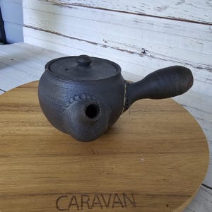 May include: A dark charcoal-colored ceramic teapot with a side-mounted handle and a matching circular lid. The teapot features a textured, matte finish and a small, rounded spout with decorative embossed detailing around the base of the spout.