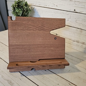 May include: A dark brown wooden tablet or phone stand featuring a rectangular back panel with a cutout notch on the top right and a horizontal ledge at the base with a recessed groove for holding a device upright in a stable position.
