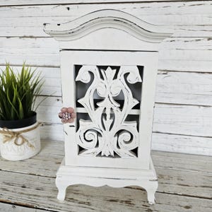 May include: White wooden cabinet with a carved design on the door and a pink flower-shaped knob. The cabinet has a distressed finish and sits on four legs.