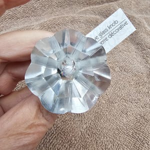 May include: A clear, faceted glass knob shaped like a flower with multiple petals. A small white tag is attached to the side of the knob, featuring the printed text 'decorative glass knob' and 'verre décorative'.