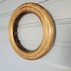 May include: A round, wooden frame with a light brown finish. The frame has a smooth, polished surface and a decorative groove around the inner and outer edges. A metal hinge is visible on the side. The frame is mounted on a light gray wall.