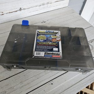 May include: A clear, rectangular Flambeau tackle box with a blue latch. The box has a label that reads "Zerust Infused" and "Extreme Environment". The label also states "Anti-Corrosion Protection".