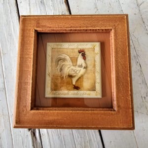 May include: Square framed artwork featuring a rooster illustration. The rooster is white with red accents, set against a beige background. The frame is a warm, reddish-brown color. Text is visible at the bottom of the artwork.