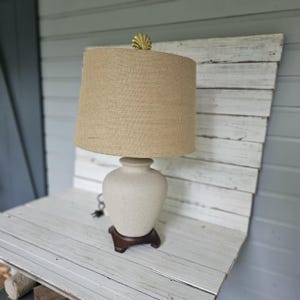 May include: A beige table lamp with a textured, off-white ceramic base and a burlap shade. The lamp has a dark brown wooden base and a gold-colored shell-shaped finial. The lamp is designed for indoor use.