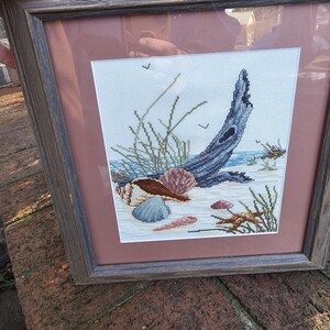 May include: Framed cross-stitch art depicting a beach scene. The artwork features a piece of driftwood, seashells, and sea grass on a sandy shore. The frame is a weathered wood color, and the matting is a muted rose color.