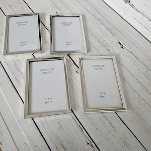 May include: Four silver-toned photo frames, each with a rectangular shape and a simple, elegant design. The frames are labeled "GODINGER SILVER" and sized for 4x6 photos. The frames are arranged on a white wooden surface.
