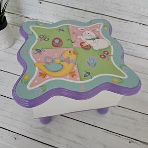 May include: A decorative Easter-themed wooden table with a scalloped edge. The top features a pastel color scheme with a bunny, a yellow bird, butterflies, and Easter eggs. The table has a white base and purple legs.