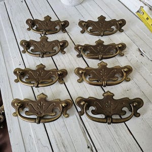 May include: Eight antique brass drawer pulls with a decorative floral design. The pulls have a curved handle and are mounted on a white wooden surface. The hardware is a warm, aged brass color.