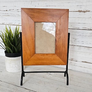 May include: A rectangular wooden picture frame with a natural wood finish. The frame is supported by a black metal stand. The frame is empty, ready for a photo. A small potted plant is visible in the background.