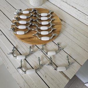 May include: A collection of cabinet pulls with a silver finish and white ceramic center. The pulls are arranged on a wooden surface and also displayed separately. The hardware is designed for furniture and home decor projects.