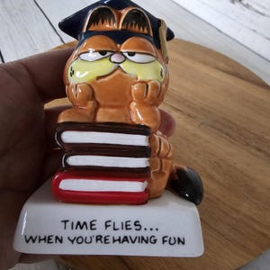 May include: A ceramic Garfield figurine wearing a graduation cap, resting on a stack of books. The orange cat is depicted with a grumpy expression. The base reads "TIME FLIES... WHEN YOU'RE HAVING FUN."
