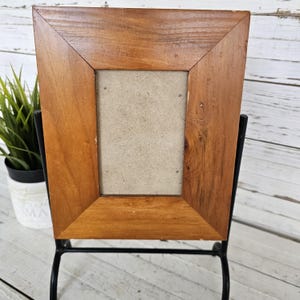 May include: A rectangular wooden picture frame with a light brown finish. The frame has a beveled edge and a neutral-colored backing. It is displayed on a black metal stand, with a small potted plant visible in the background.