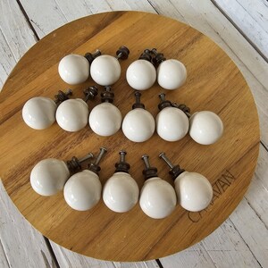 May include: A collection of white ceramic cabinet knobs with dark metal hardware. The knobs are arranged on a round wooden cutting board. The knobs have a smooth, glossy finish and are ready to be installed on furniture.
