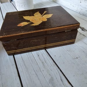 May include: A rectangular wooden box with a dark brown finish. The lid features a decorative floral inlay in shades of yellow and orange. The box has two thin, parallel gold-colored stripes around the middle.