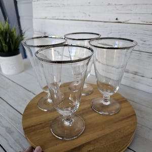 May include: Four clear glass goblets with silver rims and stems are arranged on a round wooden tray. The goblets have a classic, elegant design, perfect for serving beverages. The background is a light-colored wooden surface.