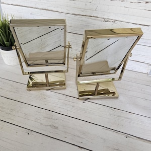 May include: Two gold-toned, rectangular vanity mirrors. Each mirror has a square base and a pivoting frame. The mirrors are set against a light-colored, wooden background. The mirrors are designed for tabletop use.