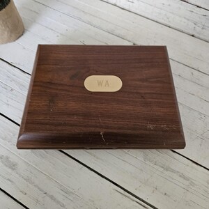 May include: A rectangular wooden box with a dark brown finish. A small, oval-shaped brass plate with the initials "WA" is centered on the lid. The box is set against a white wooden surface.