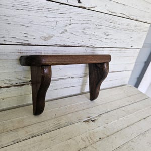May include: A small, dark brown wooden wall shelf featuring a smooth, rectangular top surface supported by two curved, decorative brackets. The wood grain is visible throughout the piece, showcasing a deep, rich stain and a polished finish.