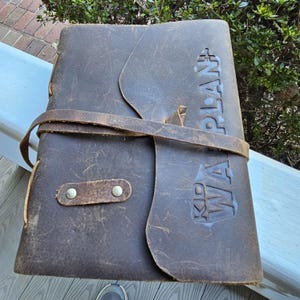 May include: A dark brown leather journal with the words "KID WA PLAN" embossed on the cover. The journal is secured with a leather strap and a small, riveted leather tab closure. The leather has a distressed appearance.