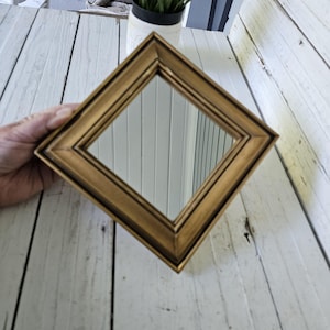 May include: A gold-colored, diamond-shaped mirror with a decorative frame. The mirror is held in a hand, showcasing its reflective surface. The frame has a layered design, adding depth to the piece. The mirror is a decorative item.