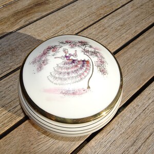 May include: A white ceramic trinket box with a gold rim and a painted design of a woman in a pink and white dress standing under a tree.