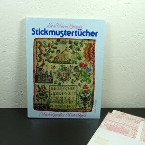 Könnte beinhalten: Ein Buch mit dem Titel "Stickmustertücher" von Eva-Maria Leszner, mit einem gestickten Design auf dem Umschlag. Das Cover-Design umfasst florale und architektonische Elemente mit deutschem Text. Mehrere Blätter Millimeterpapier sind sichtbar.