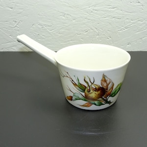May include: A white ceramic ladle with a long handle. The ladle's exterior features a botanical design with brown, green, and yellow accents. The ladle is designed for serving soups or sauces.
