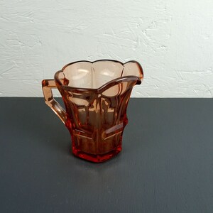 May include: A pink glass creamer with a fluted rim and a geometric design. The creamer has a handle and is sitting on a dark gray surface.