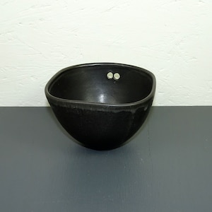 Puede incluir: Un cuenco de cerámica negro con forma cuadrada y bordes redondeados. El cuenco tiene dos pequeños agujeros en la parte superior.