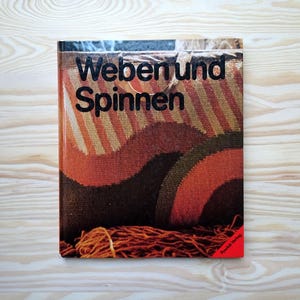 Può includere: Un libro intitolato "Weben und Spinnen" con una copertina colorata con un disegno tessuto nei toni dell'arancione, marrone e beige. Un fascio di filo marrone è in basso. Il libro è su una superficie di legno chiaro.