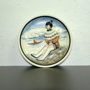 May include: A ceramic plate with a raised image of a person wearing a colorful sweater and sitting on a rock. The person is looking to the left and there is a small boat in the water behind them. The plate has a blue and white border.
