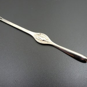 May include: A silver-toned, long, thin, and pointed tool with a decorative design in the middle. The tool is used for removing the core from apples.