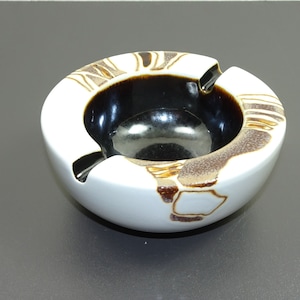 Può includere: Posacenere in ceramica bianca con smalto marrone e nero. Il posacenere è rotondo e ha una ciotola larga e poco profonda.