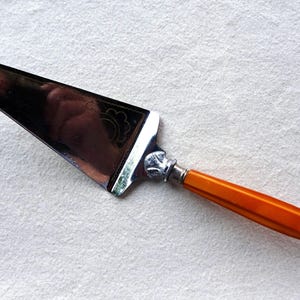 Könnte beinhalten: Ein silberner Kuchenschieber mit einem orangefarbenen Kunststoffgriff. Der Schieber hat ein dekoratives Design auf der Klinge.