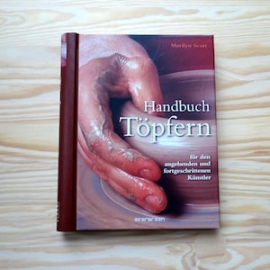 Pode incluir: Um livro intitulado "Handbuch Töpfern" com uma capa que mostra mãos trabalhando com argila. O título está em branco. A lombada do livro é marrom. O nome da autora, Marilyn Scott, e o logotipo Evergreen são visíveis.
