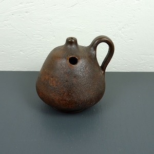 Puede incluir: Una lámpara de aceite de cerámica marrón con asa y un agujero en la parte superior. La lámpara tiene un acabado rústico y texturizado.