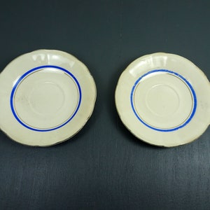 May include: Two white ceramic saucers with a blue and gold trim. The saucers are on a dark gray background.