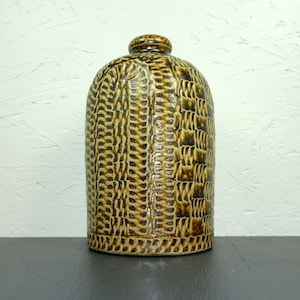 May include: A brown ceramic bottle with a textured, woven pattern. The bottle has a rounded body and a narrow neck.