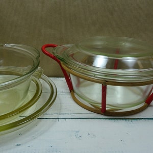 Op de afbeelding: Twee glazen ovenschaaltjes met deksels. De grotere schaal heeft een rood en bruin gevlochten handvat. De kleinere schaal is gestapeld op een heldere glazen plaat.