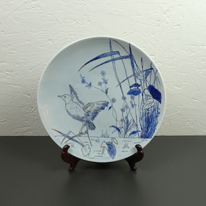 May include: A white ceramic plate with a blue and gold design of a bird and flowers. The plate is on a wooden stand.